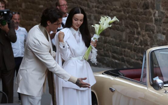 Damiano David in doppiopetto color panna apre la portiera della Lancia d'epoca color crema a Dove Cameron, in tailleur bianco con un mazzo di calle, all'arrivo al Palazzo Comunale di Montalcino