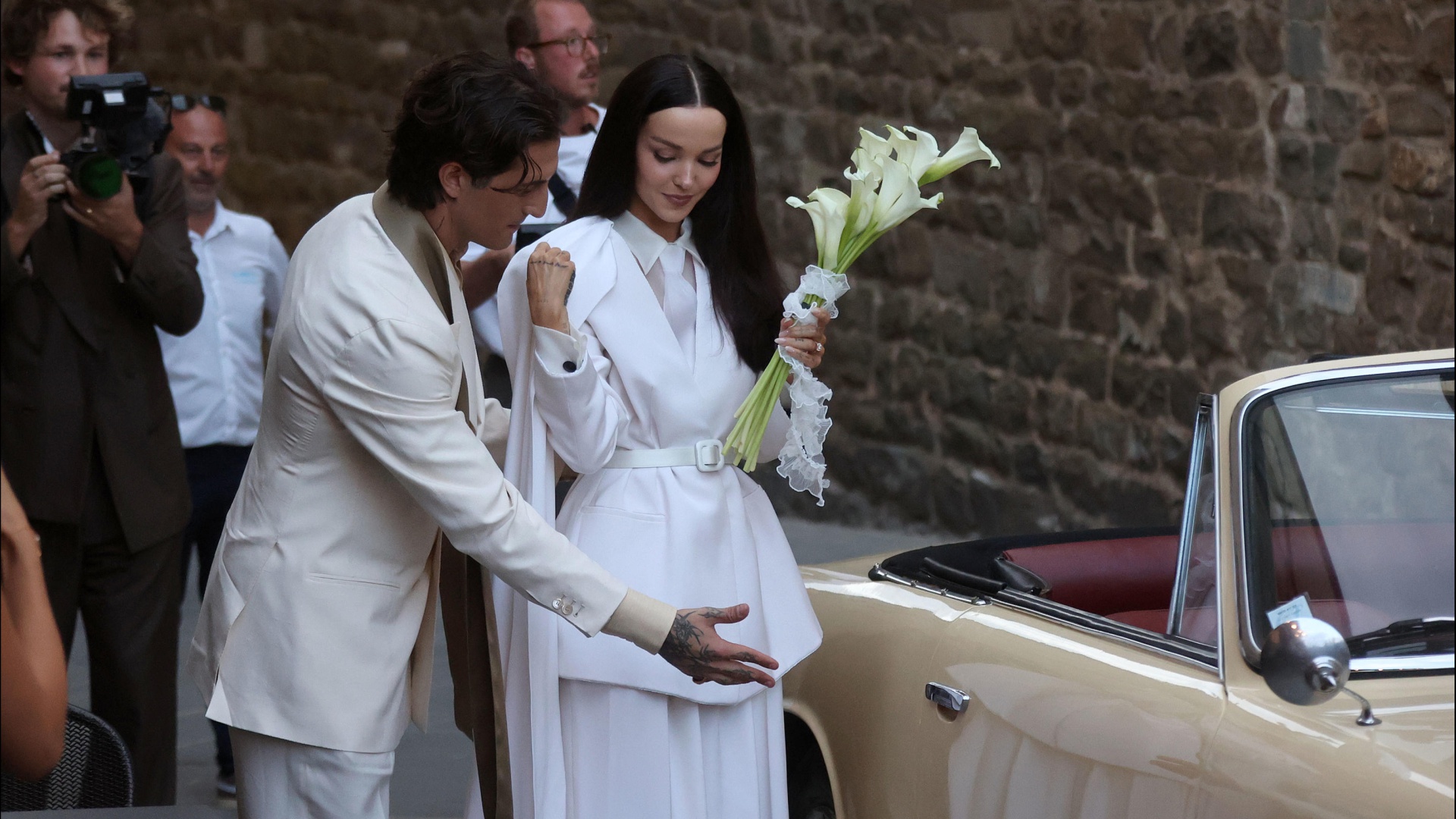 Damiano David in doppiopetto color panna apre la portiera della Lancia d'epoca color crema a Dove Cameron, in tailleur bianco con un mazzo di calle, all'arrivo al Palazzo Comunale di Montalcino