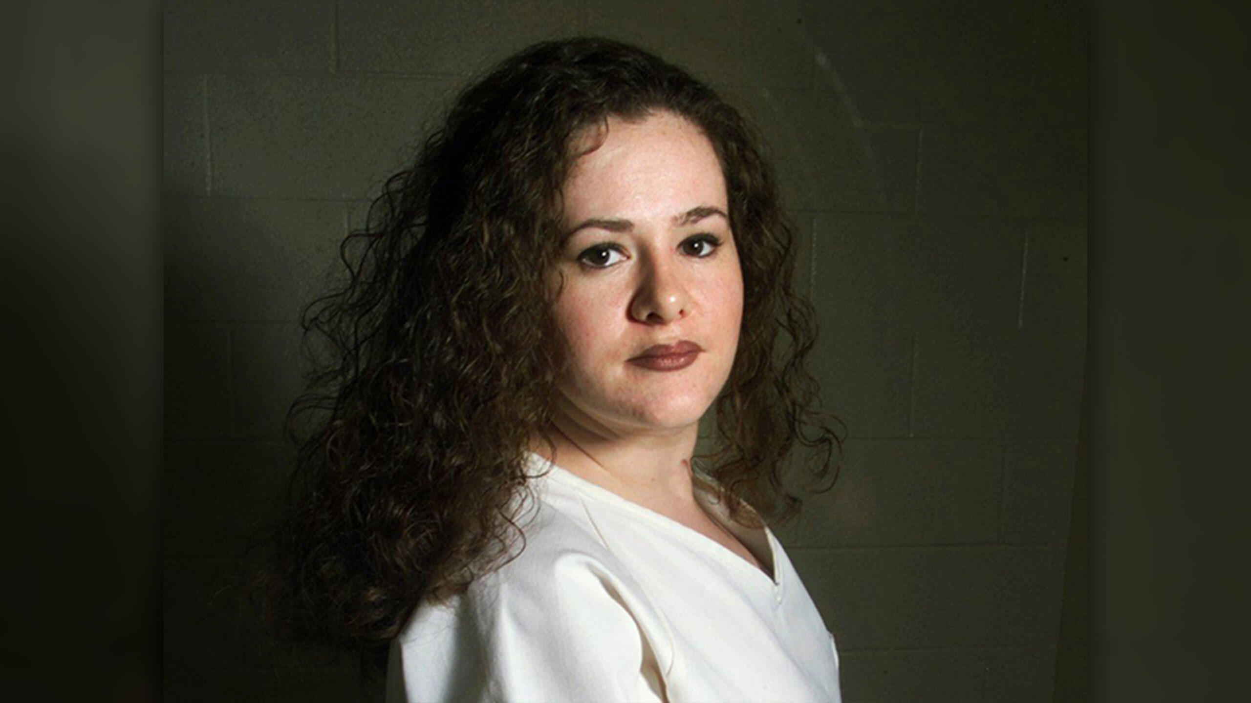 Ritratto di Christa Pike: capelli ricci scuri, maglia bianca, davanti a un muro di blocchi di cemento.